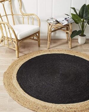 Braided round jute rug features a black center and natural tan border, placed in a room with a small table, houseplant, and rattan chair.