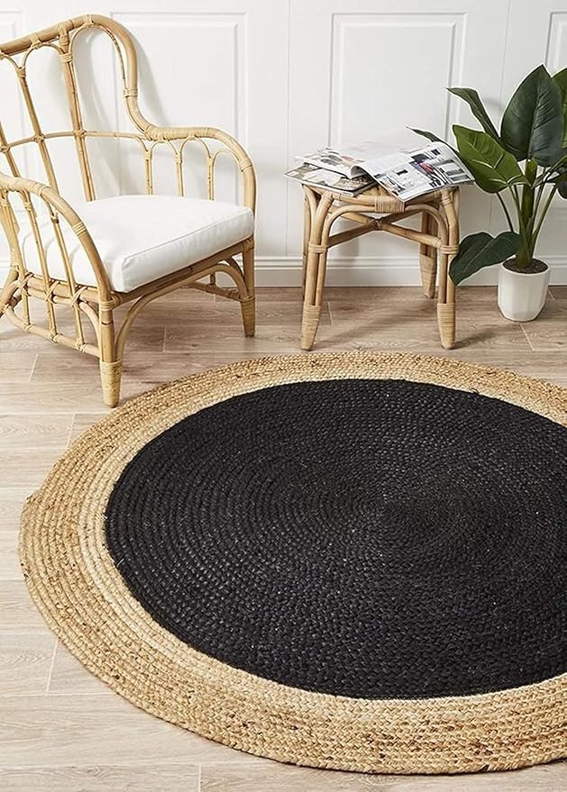 Braided round jute rug features a black center and natural tan border, placed in a room with a small table, houseplant, and rattan chair.