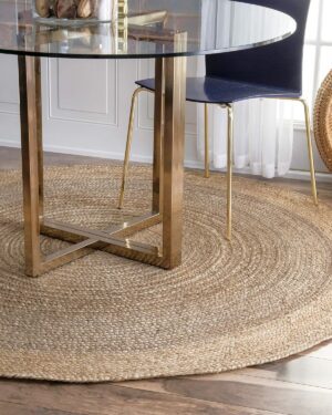 Round jute rug sits beneath a glass-topped table with gold legs and a modern chair on a wood floor near a wicker chair.