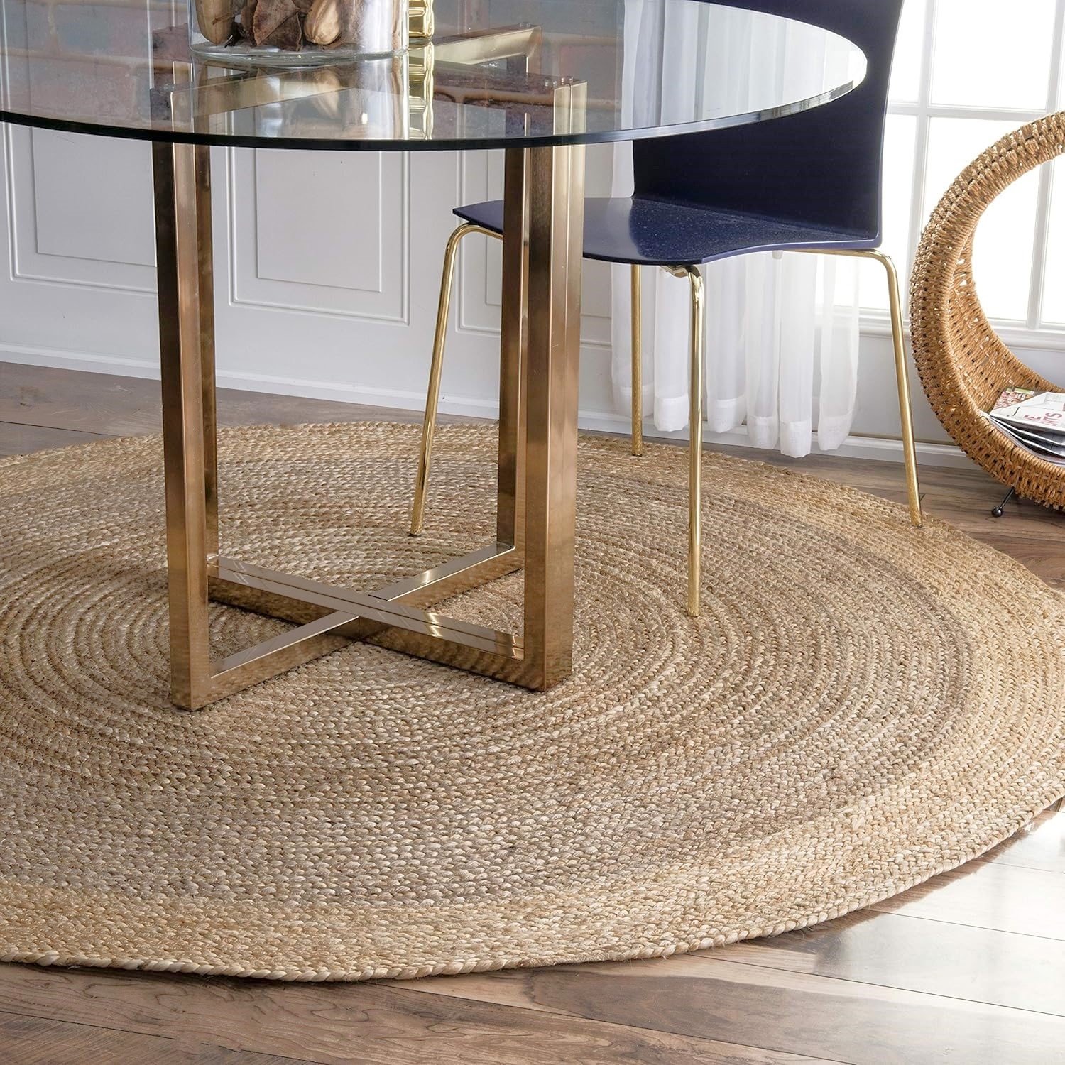 Round jute rug sits beneath a glass-topped table with gold legs and a modern chair on a wood floor near a wicker chair.